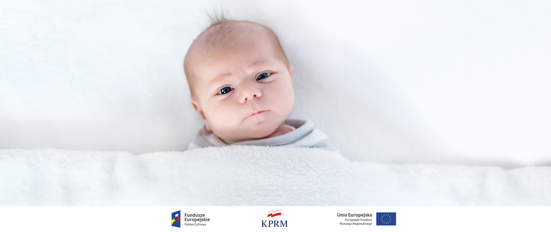 Ubrany na biało niemowlak leżący na białym łóżku pod białą kołdrą - szeroki plan, widać tylko głowę i ramiona dziecka.