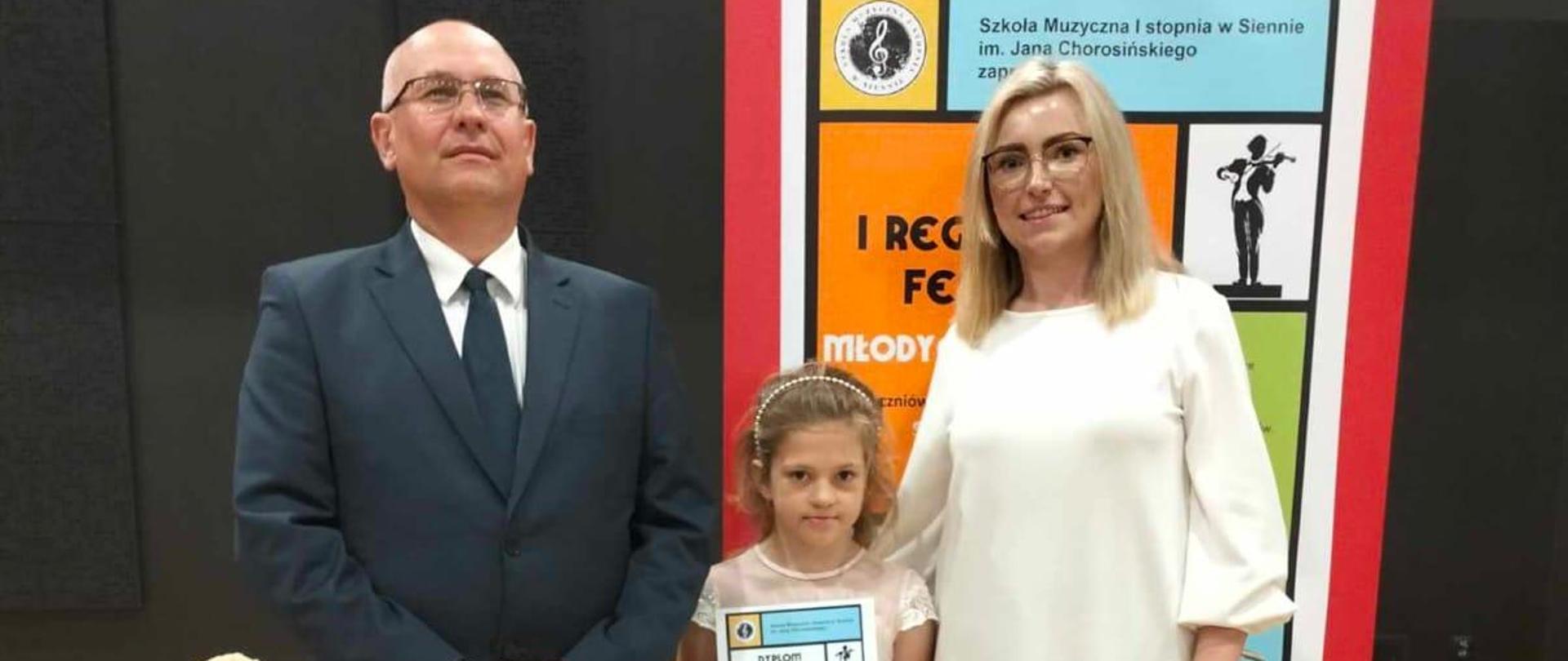 Na zdjęciu widać po prawej stronie panią profesor która przygotowywała uczennicę na konkurs na środku zdjęcia uczennica z dyplomem a po lewej stronie pan akompaniator w garniturze w tle widoczny wazon z kwiatami po lewej stronie a za uczennicą widoczny plakat który reklamował konkurs.