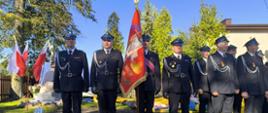 Na zdjęciu kilku strażaków OSP w mundurach galowych. Stoją w szeregu po lewej stronie zdjęcia i w dwuszeregu po prawej stronie zdjęcia. Jeden strażak w środku zdjęcia trzyma sztandar. W tle widać trzy flagi polski, fragment budynku i drzewo. Powyżej bezchmurne niebieskie niebo.