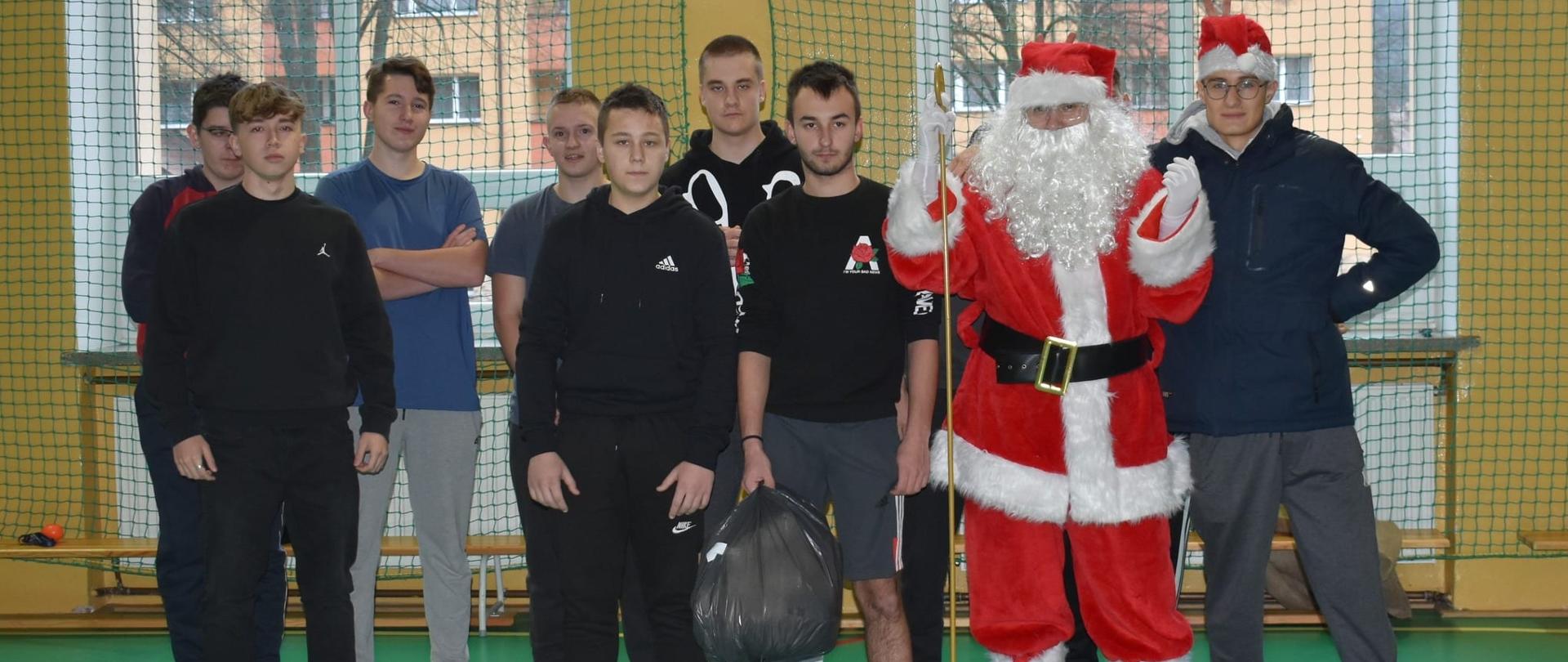 Zdjęcie grupowe na sali gimnastycznej. Święty Mikołaj i jego pomocnik w mikołajkowej czapce pozują z grupą młodych mężczyzn.