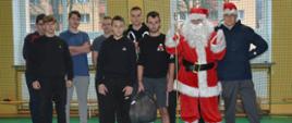 Zdjęcie grupowe na sali gimnastycznej. Święty Mikołaj i jego pomocnik w mikołajkowej czapce pozują z grupą młodych mężczyzn.
