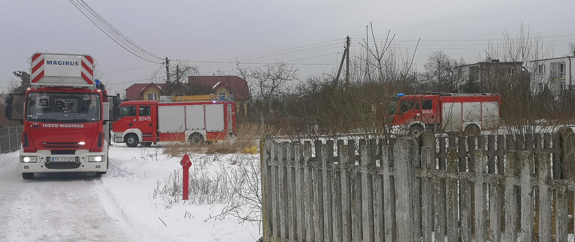 Zdjęcie przedstawia trzy samochody pożarnicze ustawione przed posesją, na której doszło do pożaru. Po prawej stronie widać betonowy płot oraz hydrant. Droga dojazdowa jak również cała okolica pokryta jest śniegiem.