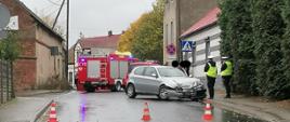 Widać samochód osobowy, który jest na drodze przy chodniku, w pobliżu dwóch policjantów, z tyłu samochód strażacki
