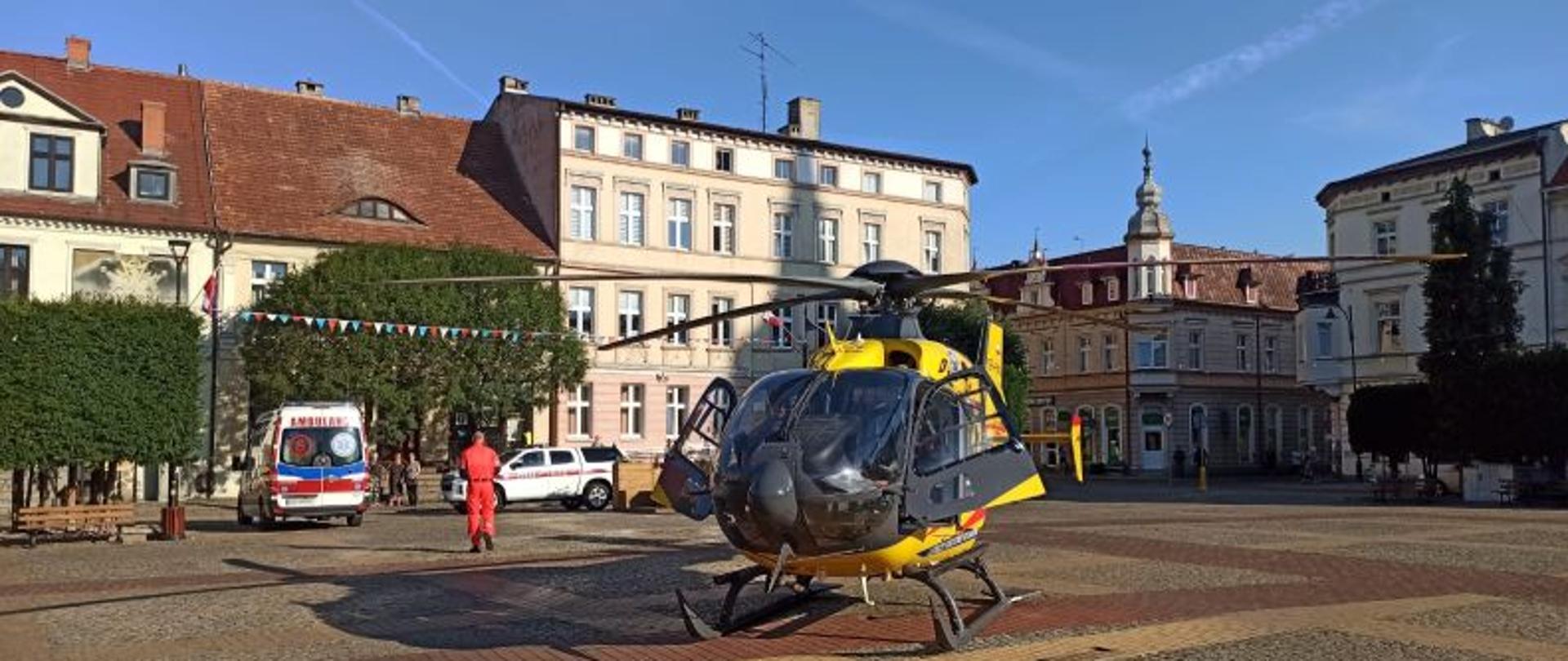 Centralny punkt zdjęcia stanowi helikopter . Helikopter koloru żółtego. Stoi na środku placu w centrum miasta. W oddali kościół a po bokach kamienice. Tło stanowi niebieski niebo.