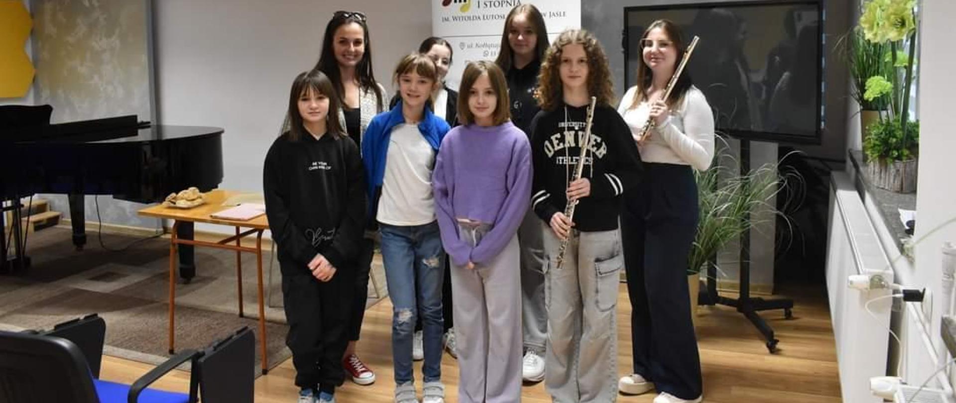 Na zdjęciu widać uczniów klasy fletu biorących udział w warsztatach, nauczyciela fletu oraz prowadzącą warsztaty, dwie osoby trzymają w ręku flet poprzeczny, za nimi w tle umieszczony jest rollup z logo Państwowej Szkoły Muzycznej 1 stopnia w Jaśle, całość utrzymana jest w kolorystyce brązów i szarości.