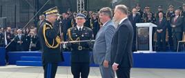 Pomorski komendant wojewódzki Państwowej Straży Pożarnej w obecności prezydenta Rzeczypospolitej Polskiej, sekretarza stanu w Ministerstwie Spraw Wewnętrznych i Administracji oraz innego strażaka całuje szablę. Przed nimi jest trybuna honorowa z zaproszonymi gośćmi. 