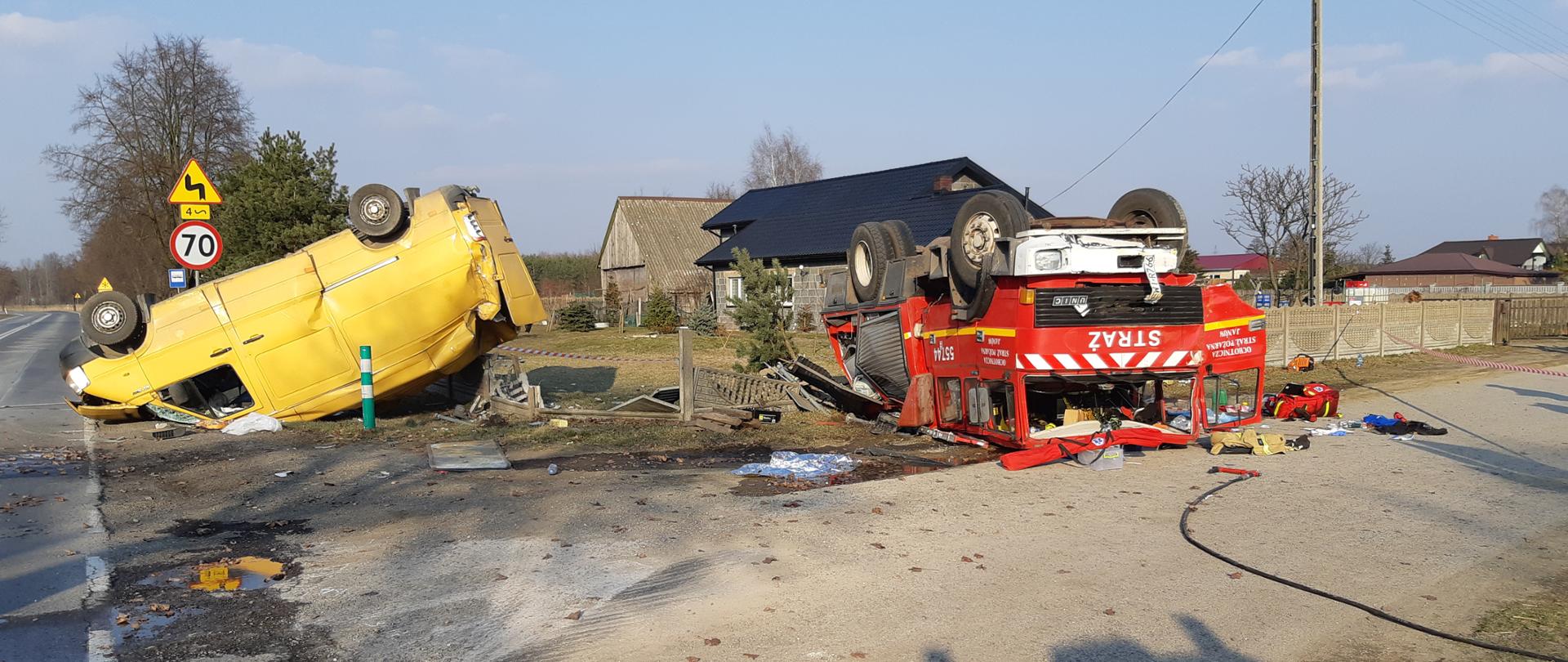 Zdjęcie przedstawia wypadek samochodu pożarniczego OSP Janów, samochód leży na dachu, obok również na dachu leży samochód dostawczy koloru żółtego, widać uszkodzone ogrodzenie posesji oraz porozrzucane elementy wyposażenia samochodu pożarniczego