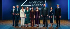 Na tle granatowej ściany z napisem 25 for Women in Science stoi czterech mężczyzn w garniturach i pięć kobiet w eleganckich sukniach, trzeci od prawej stoi wiceminister Szeptycki.