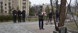 Na dworze, wiceminister Szeptycki składa wieniec biało-czerwonych kwiatów pod drewnianym krzyżem.