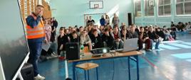 Zdjęcie przedstawia spotkanie informacyjno‑edukacyjne w sali gimnastycznej. Widoczny jest ekran projekcyjny, projektor oraz uczestnicy siedzący naprzeciw osoby prowadzącej prezentację
