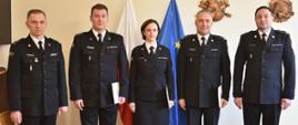 na zdjęciu pięcioro strażaków w mundurach wyjściowych, czterech mężczyzn oraz jednak kobieta, stoją w rzędzie w sali, za nimi flagi Polski i Unii Europejskiej
