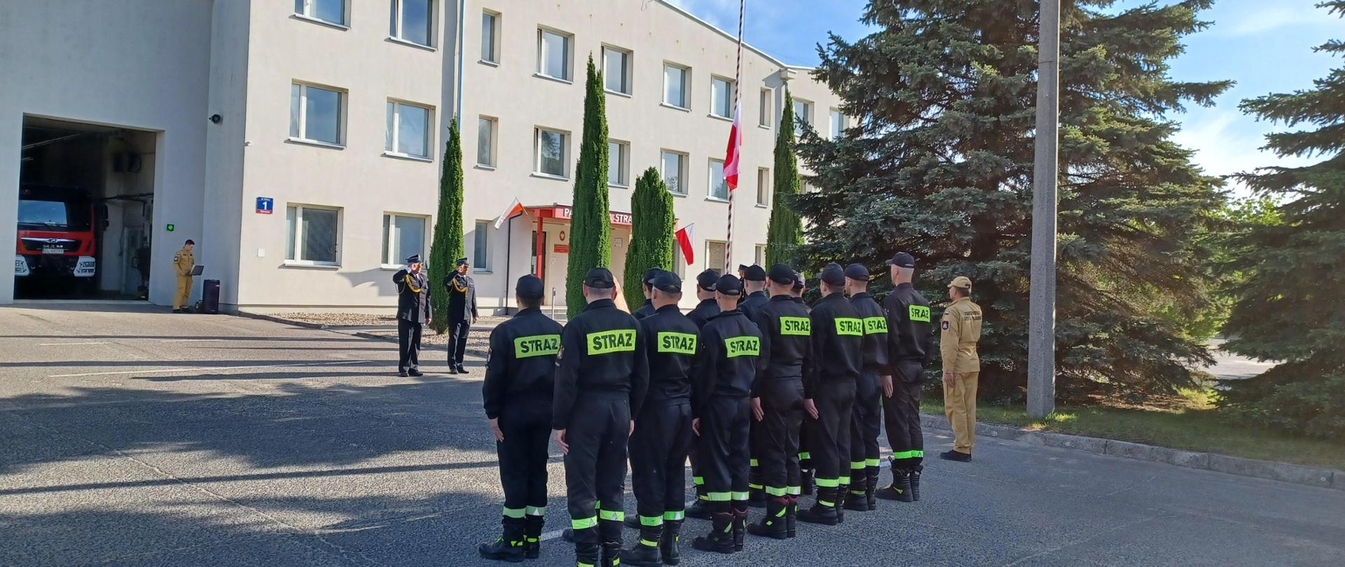 Pododdział strażaków w umundurowaniu koszarowym, dowódca i dwóch komendantów zalutują w stronę podnoszonej na maszcie biało czerwonej flagi. W tle budynek Komendy Powiatowej PSP w Opocznie. 