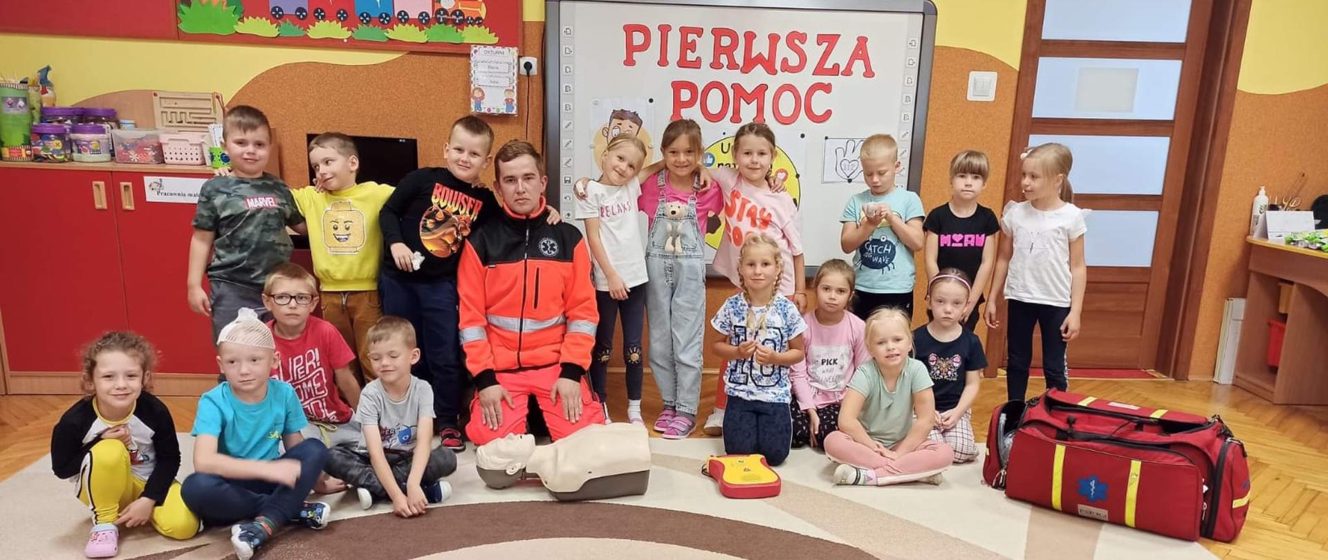 Wizyta w Przedszkolu nr 2 w Sokołowie Podlaskim - na zdjęciu na sali na dywanie strażak w mundurze ratownika klęczy w gronie przedszkolaków. W tle tablica multimedialna .