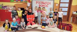 Wizyta w Przedszkolu nr 2 w Sokołowie Podlaskim - na zdjęciu na sali na dywanie strażak w mundurze ratownika klęczy w gronie przedszkolaków. W tle tablica multimedialna .
