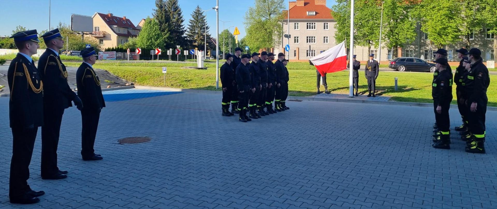 Na przeciwko siebie stoją w dwuszeregu strażacy w ubraniu koszarowym, po środku nich znajduje się maszt flagowy i poczet flagowy składający się z trzech strażaków ubranych w mundury wyjściowe. Strażacy prezentują flagę Rzeczypospolitej Polskiej.