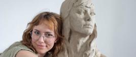 zdjęcie przedstawia młodą kobietę o falowanych, jasnobrązowych włosach i w okularach w metalowych oprawkach, czule przytulającą dużą, surową rzeźbę z gliny. Kobieta ma na sobie oliwkowozielony top i patrzy bezpośrednio w obiektyw z delikatnym uśmiechem.