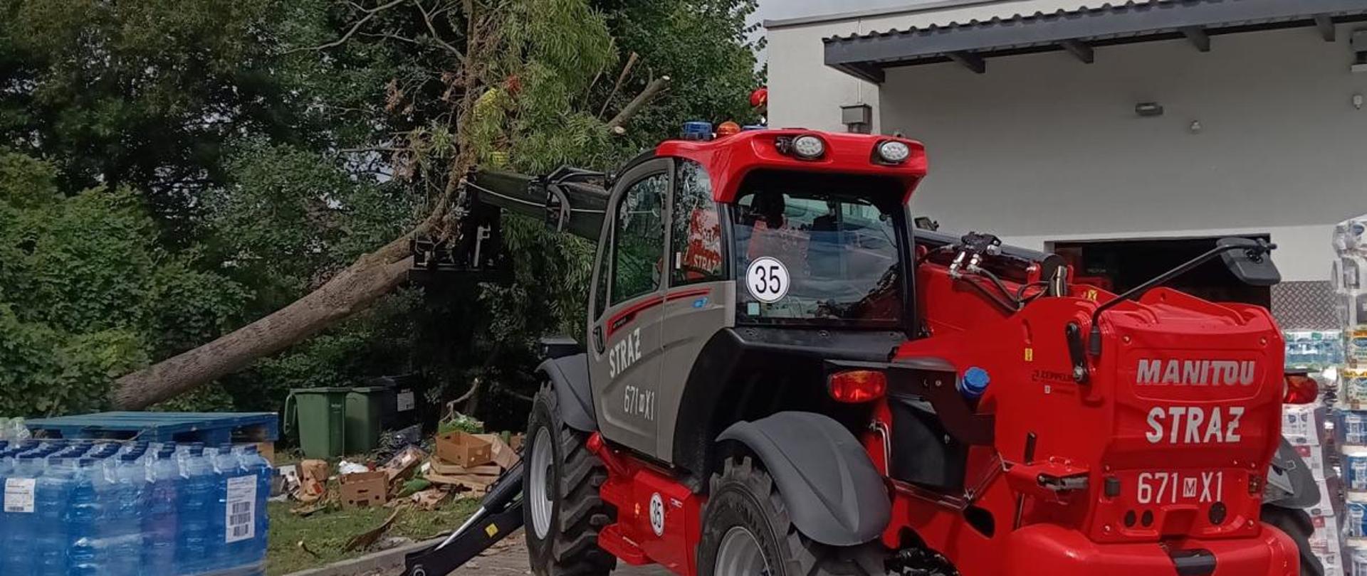 Na zdjęciu widoczna jest ładowarka teleskopowa Manitou wykorzystywana przez strażaków podczas usuwania powalonego drzewa. W tle widoczny jest uszkodzony konar oraz teren prowadzonych działań ratowniczych.
