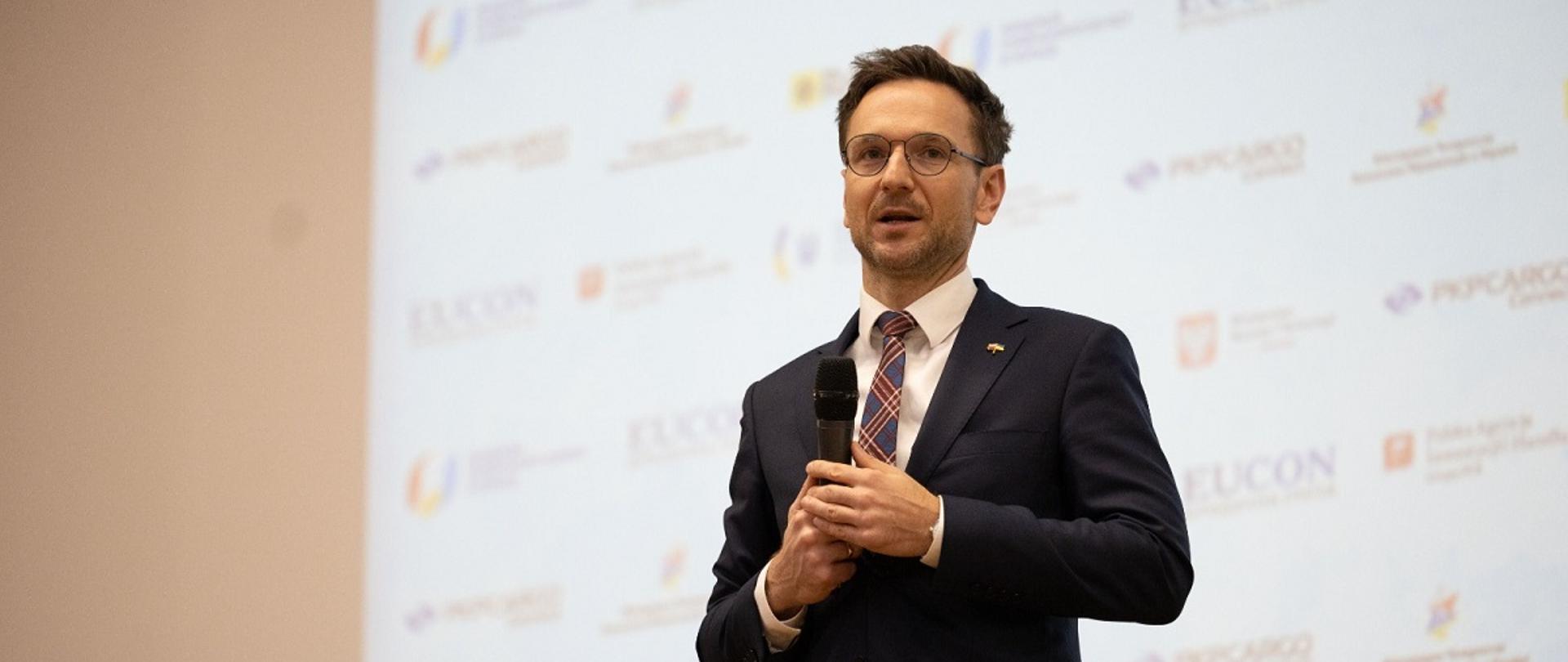 Minister rozwoju i technologii Waldemar Buda trzyma w ręku mikrofon, za jego plecami ekran wyświetlający sponsorów XV Międzynarodowego Forum „Polsko-Ukraińskie dni biznesowe”.