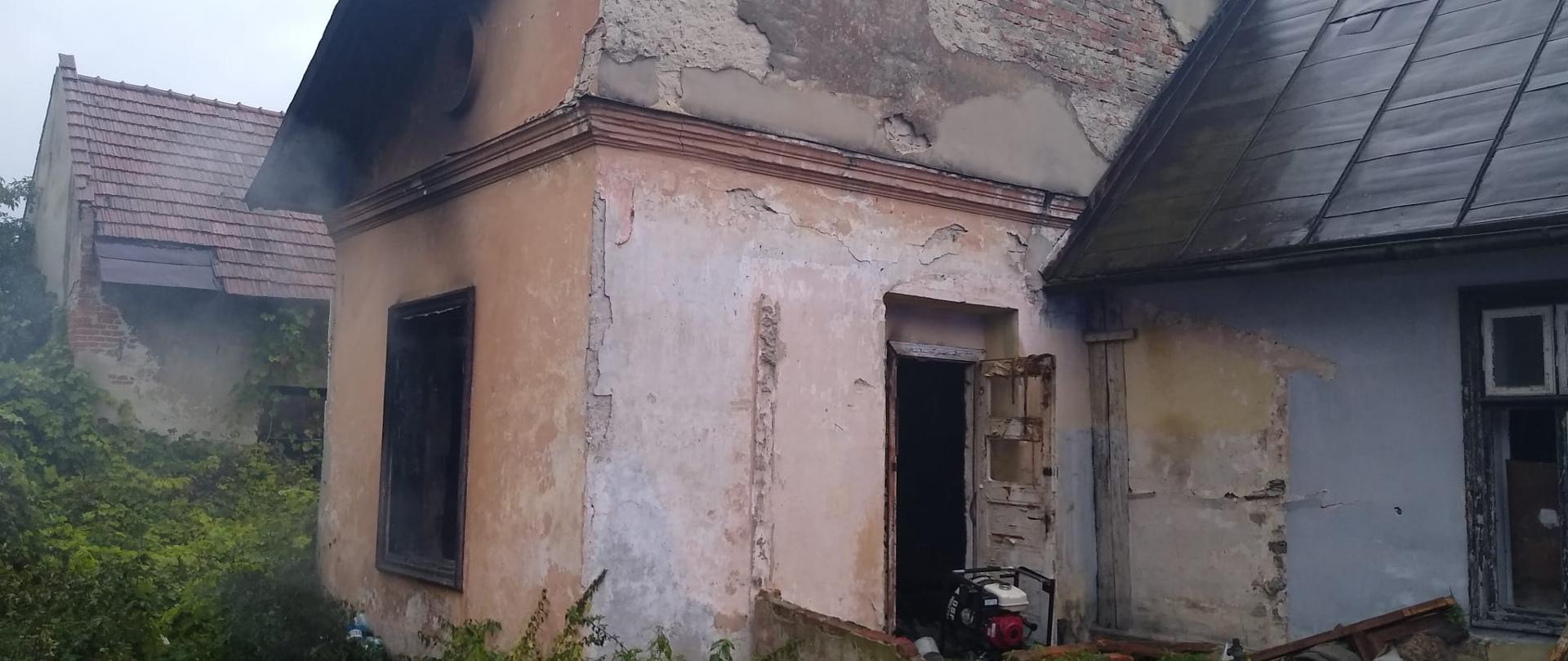 Budynek z którego wydobywa się dym. W drzwiach budynku wentylator.