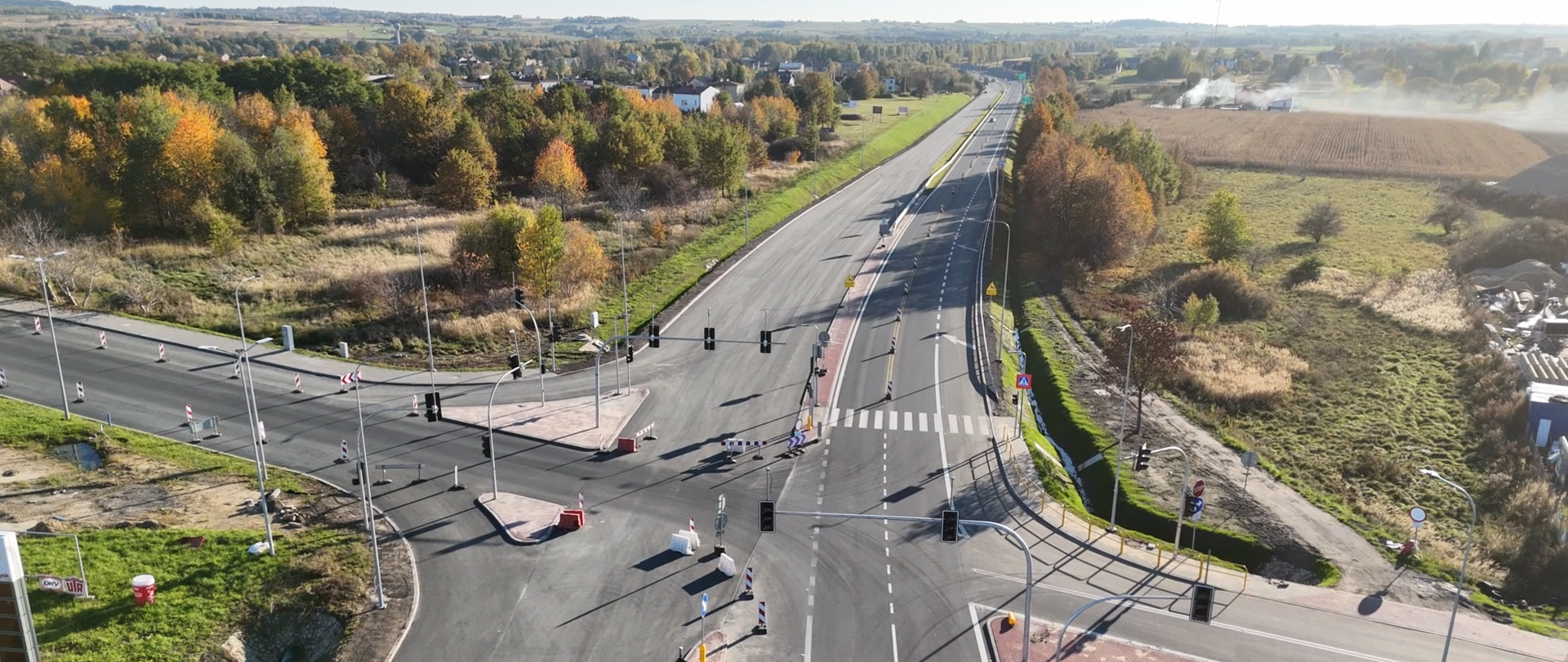 Widok z lotu ptaka na przebudowany odcinek drogi krajowej nr 91 pomiędzy Koziegłowami a Siedlcem Dużym. Na zdjęciu widać nową nawierzchnię jezdni, skrzyżowanie z sygnalizacją świetlną, oznakowane przejście dla pieszych oraz pasy ruchu. Po obu stronach drogi znajdują się tereny zielone i zabudowa rozproszona, w tle widoczny krajobraz pól i drzew w jesiennej kolorystyce.