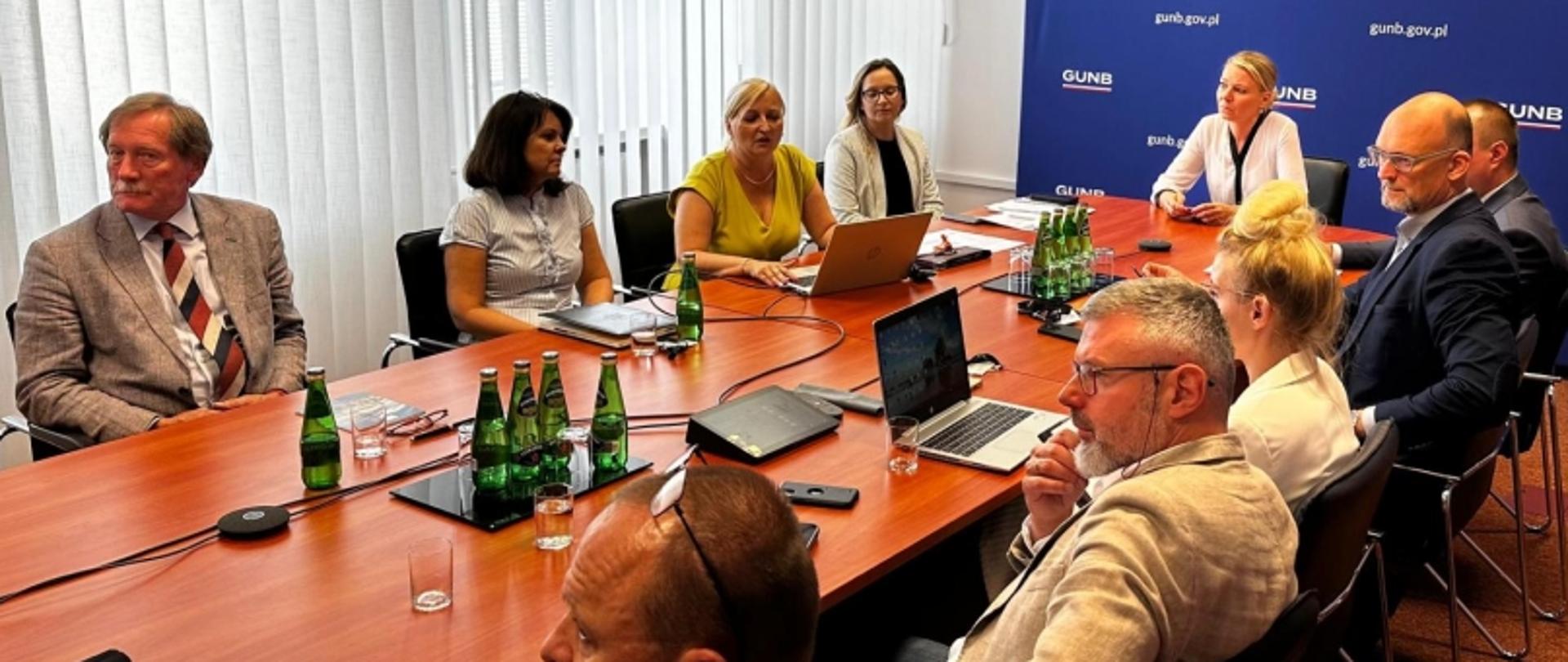 Grupa osób przy stole konferencyjnym podczas spotkania w GUNB; na stole laptopy, notatniki i butelki wody, w tle niebieska ściana z logo „GUNB” i adresem „gunb.gov.pl”.