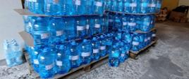 Widok z boku. Woda pitna w dużych pojemnikach składowana na paletach. 