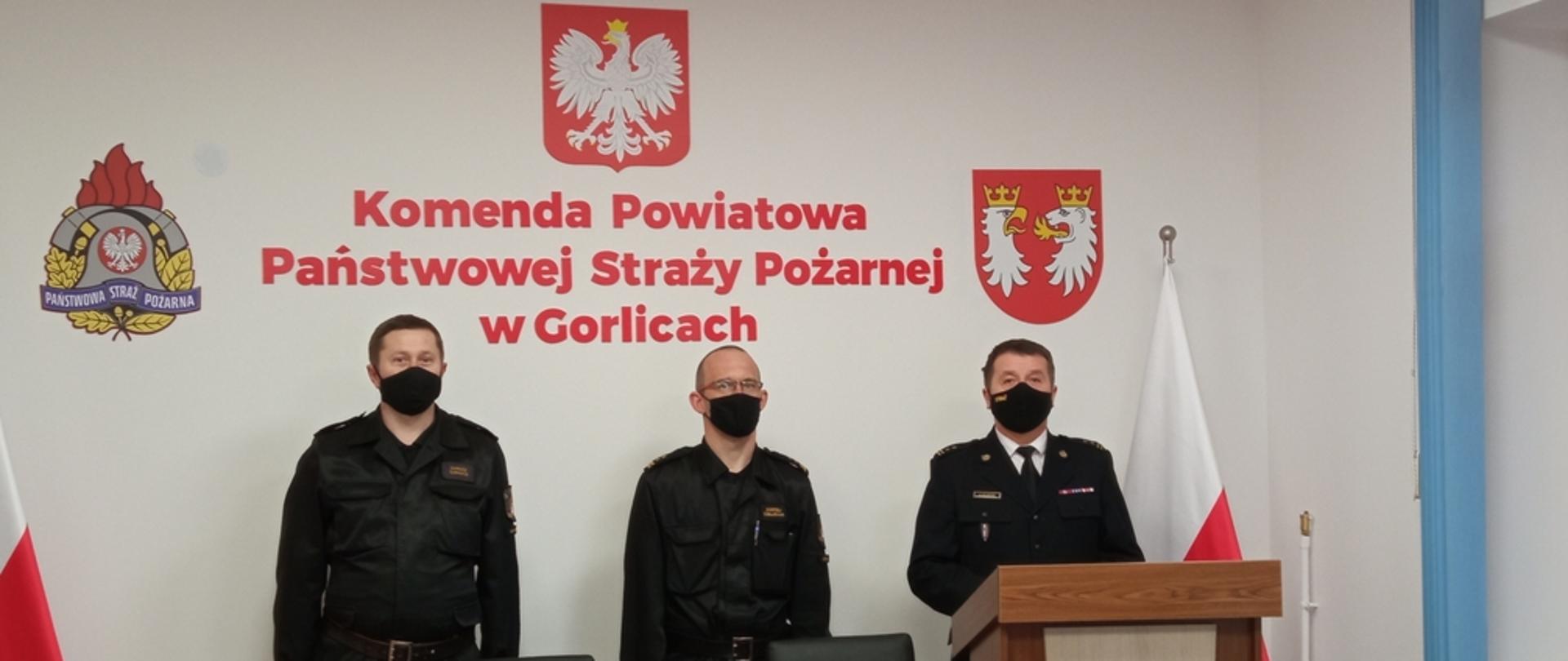 Na zdjęciu widoczny po lewej stronie w ubraniu wyjściowym Komendant Powiatowy Państwowej Straży Pożarnej w Gorlicach bryg. Krzysztof Gładysz w środku w ubraniu dowódczo sztabowym Naczelnik Wydziału ds. Operacyjno-szkoleniowych w Gorlicach po lewej stronie w ubraniu dowódczo sztabowym mł.bryg. Dariusz Surmacz Z-ca D-cy Jednostki Ratowniczo-Gaśniczej w Gorlicach. Strażacy stoją na tle białej ściany z napisem koloru czerwonego Komenda Powiatowa Państwowej Straży Pożarnej w Gorlicach po bokach flagi biało czerwone. 