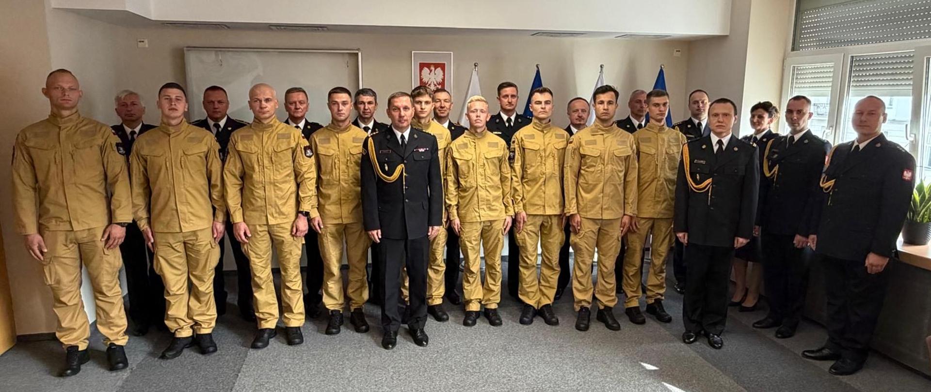 Zdjęcie grupowe strażaków w jasnym pomieszczeniu. W pierwszym rzędzie stoi grupa mężczyzn w piaskowych mundurach służbowych oraz strażacy w ciemnych mundurach galowych z sznurami galowymi. W drugim rzędzie stoją pozostali funkcjonariusze w mundurach galowych. W tle na ścianie wisi godło Polski oraz stoją flagi państwowe i urzędowe.