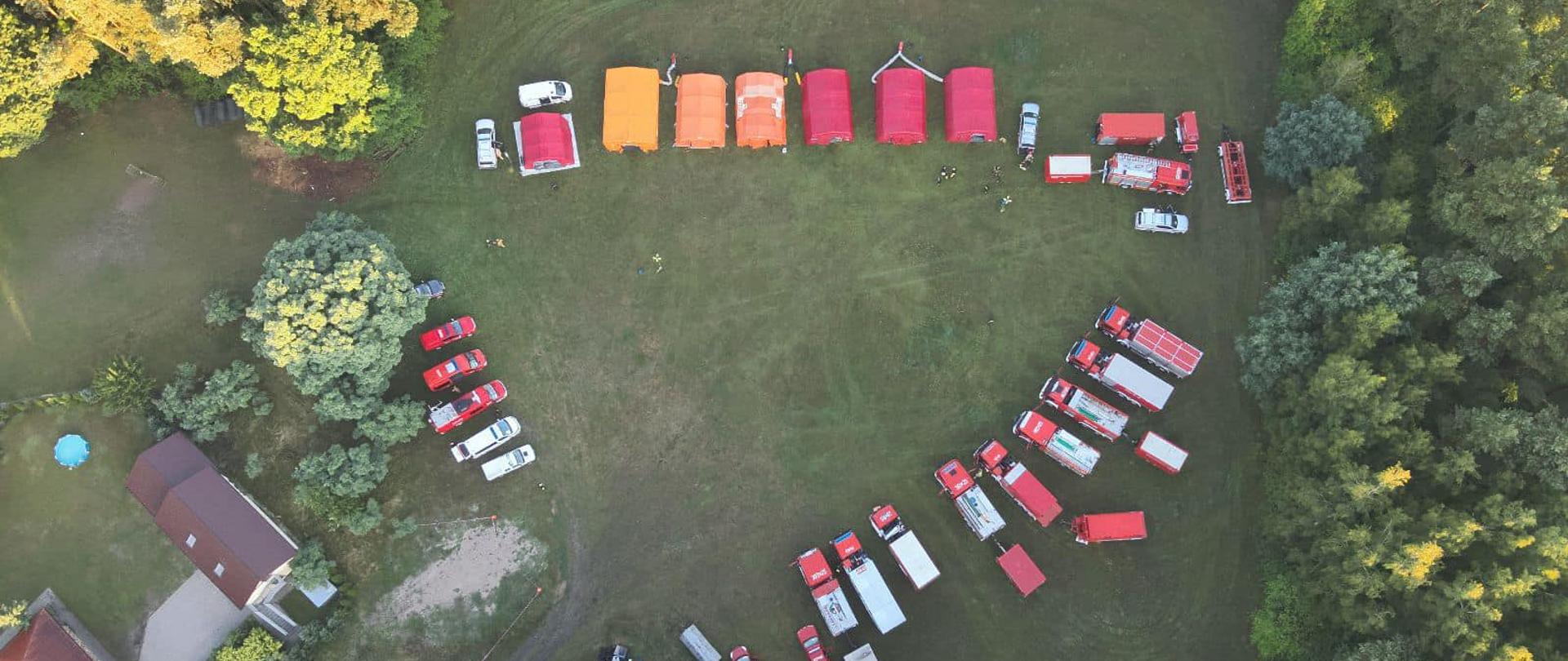 Widok z góry na teren obozowiska - kilka namiotów w szeregu, w innym szeregu ustawione pojazdy pożarnicze. Widać także budynki, a w koło drzewa i droga dojazdowa do polany.