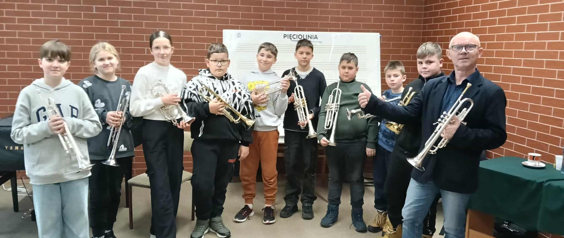 Grupa uczniów stojących z trąbkami obok prowadzącego warsztaty profesora; wszyscy pozują do wspólnego zdjęcia w sali szkolnej na tle tablicy pięciolinii i ceglanej ściany.