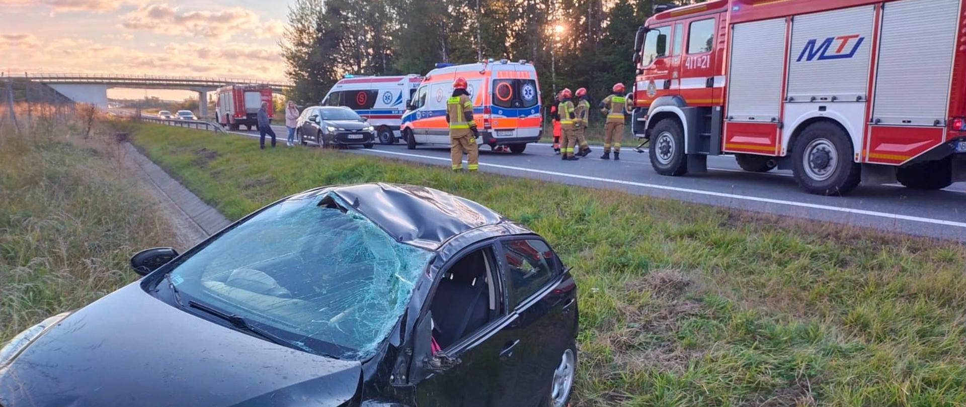 Czarny samochód osobowy w rowie na poboczu drogi. Na drodze ustawione samochody straży pożarnej i dwie karetki. W tle las.