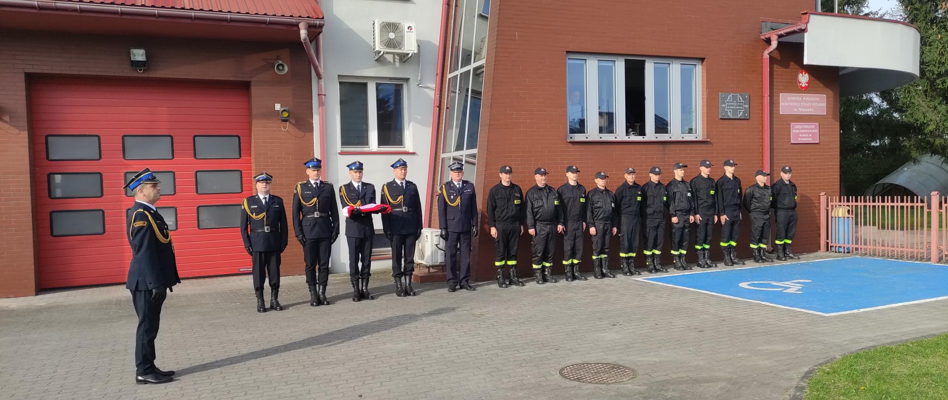 Przed budynkiem KP PSP we Włoszczowie trwa zmiana służby połączona z uroczystym podniesieniem flagi państwowej na maszt przy dźwiękach hymnu narodowego