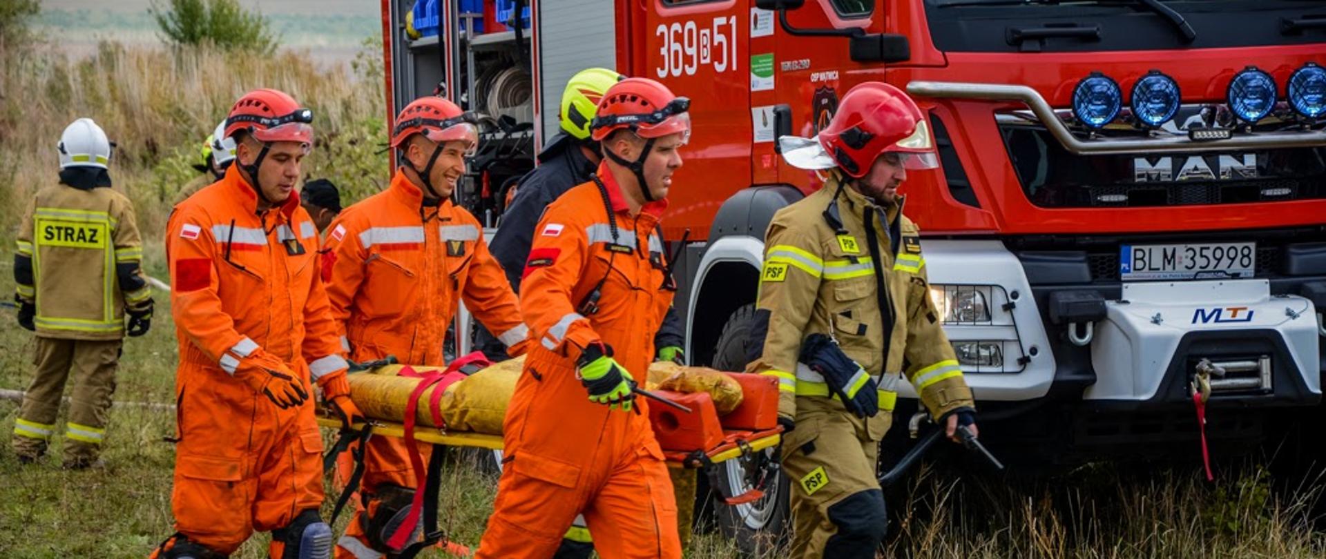 Strażacy podczas transportu osoby poszkodowanej na desce medycznej. W tle widoczny pojazd ratowniczo-gaśniczy