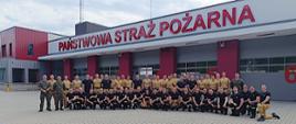 Na zdjęciu widoczna jest duża grupa strażaków – około 70 osób – ustawionych przed budynkiem straży pożarnej z napisem „PAŃSTWOWA STRAŻ POŻARNA” w dużych, czerwonych literach. Osoby na zdjęciu strażacy - uczestnicy szkolenia kompanii reprezentacyjnej – część z nich ma na sobie czarne koszulki i bojowe spodnie, a inni beżowe koszulki, a niektórzy mundury służbowe - pisakowe.
Grupa ustawiła się w dwóch rzędach: pierwszy rząd klęczy lub kuca, a drugi stoi za nimi. W tle widać nowoczesny budynek straży pożarnej w kolorach czerwonym i szarym z numerami bram wjazdowych (np. 5 i 6) namalowanymi na dużych drzwiach garażowych. 