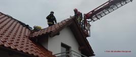 Dach budynku mieszkalnego. Z prawej strony widoczne ramię strażackiego podnośnika hydraulicznego, w koszu stoi strażak w czerwonym hełmie. Na twarzy ma maskę, a na plecach założony aparat oddechowy. Obok niego stoi na dachu podobnie ubrany strażak w białym hełmie, a po jego lewej stronie inny strażak w pozycji klęczącej. 