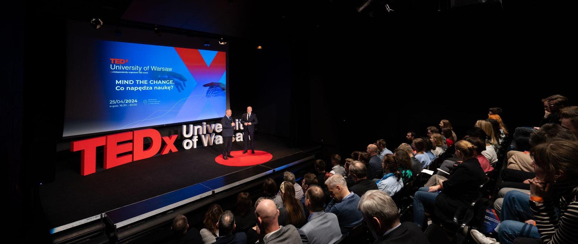 Zdjęcie z boku, zaciemniona sala wypełniona siedzącymi w rzędach krzeseł ludźmi, przed nimi na podwyższeniu stoi wiceminister Gzik i mężczyzna w garniturze, za nimi napis TEDx University of Warsaw ułożony z dużych liter.