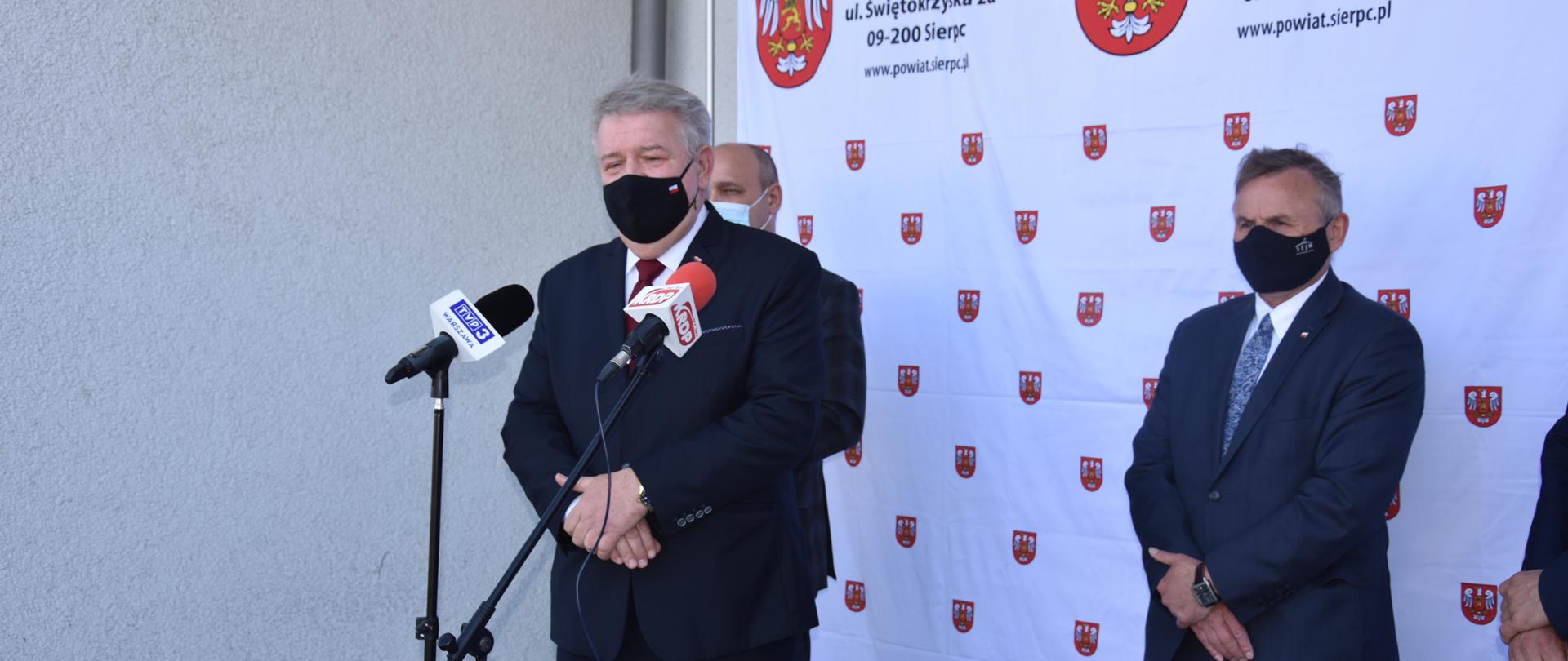 Zdjęcie przedstawia ubranego w garnitur mężczyznę, który jest w trakcie wystąpienia publicznego. W tle stoi dwóch mężczyzn w garniturach na tle ścianki białej ścianki wizerunkowej Starostwa Powiatowego w Sierpcu. 