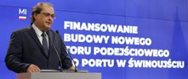 Wiceminister infrastruktury Marek Gróbarczyk stoi przy mównicy i przemawia do mikrofonów, za jego plecami ekran informujący o finansowaniu budowy nowego toru podejściowego w porcie Świnoujście