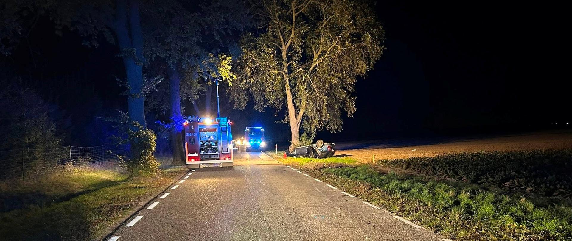 Samochody strażacki na drodze, samochód osobowy po dachowaniu w rowie blisko drzewa 