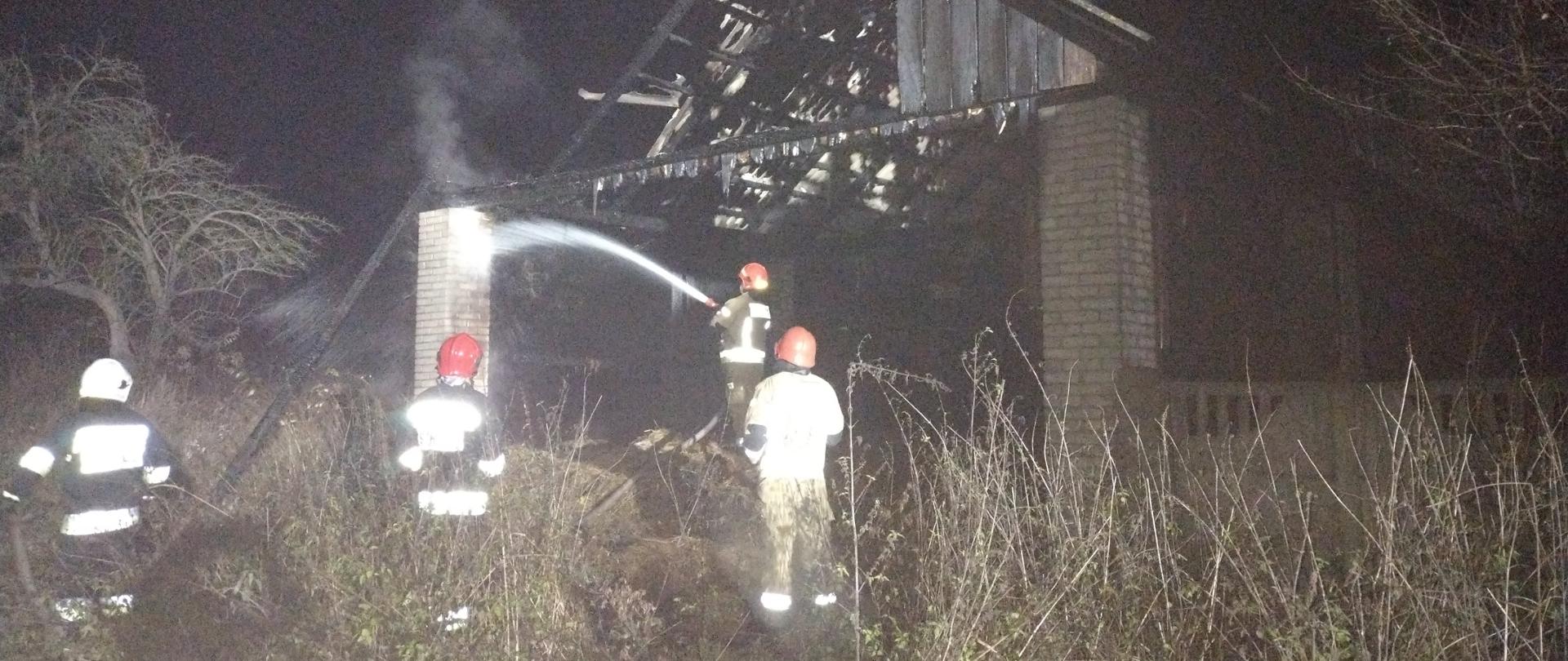 Na zdjęciu widać 4 strażaków, którzy dogaszają tlące się elementy stodoły po pożarze. Działania prowadzone są w porze nocnej.