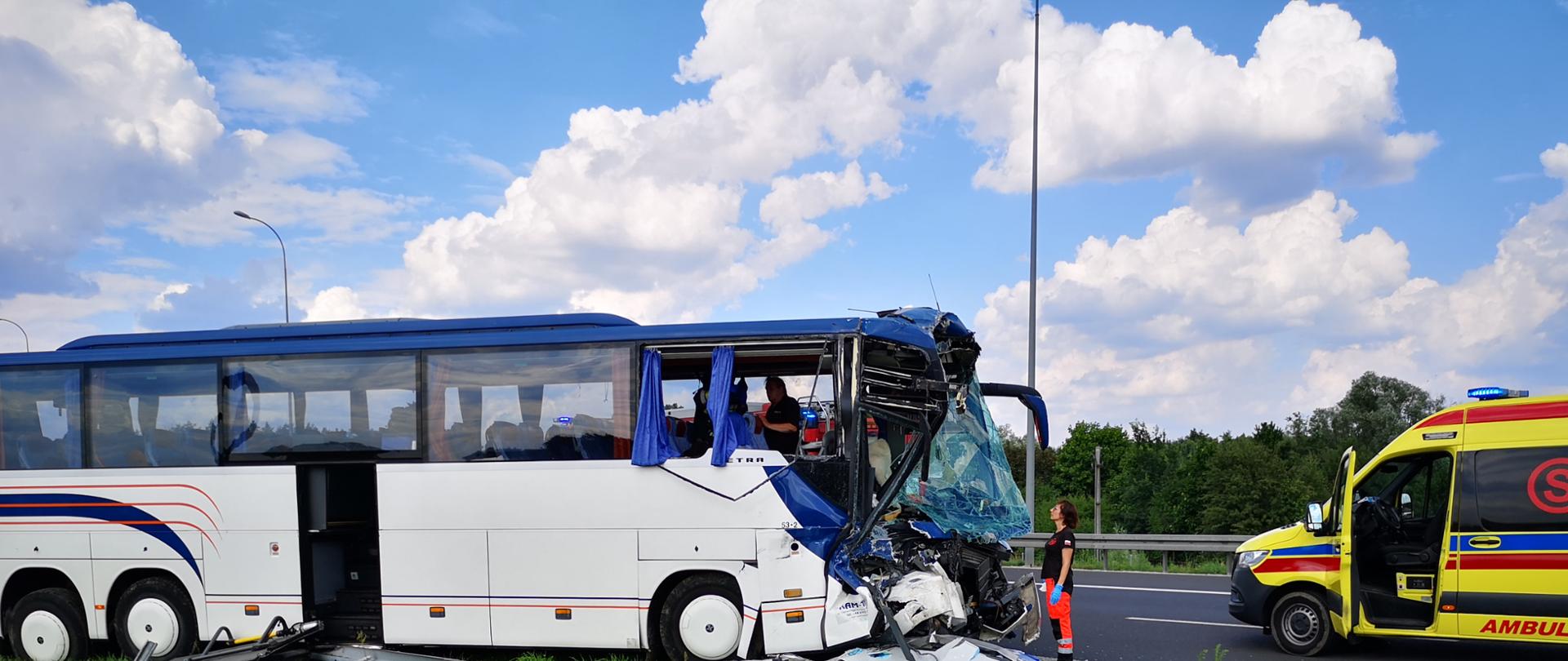 Uszkodzony autokar na wskutek wypadku . Rozbita przednia czołowa część autokaru. Przed rozbitym autokarem służby współdziałające tj. pogotowie ratunkowe i ratownik medyczny. Na pasie zieleni leżą uszkodzone barierki zabezpieczające od pasa autostrady i rozbite, potłuczone elementy autokaru.