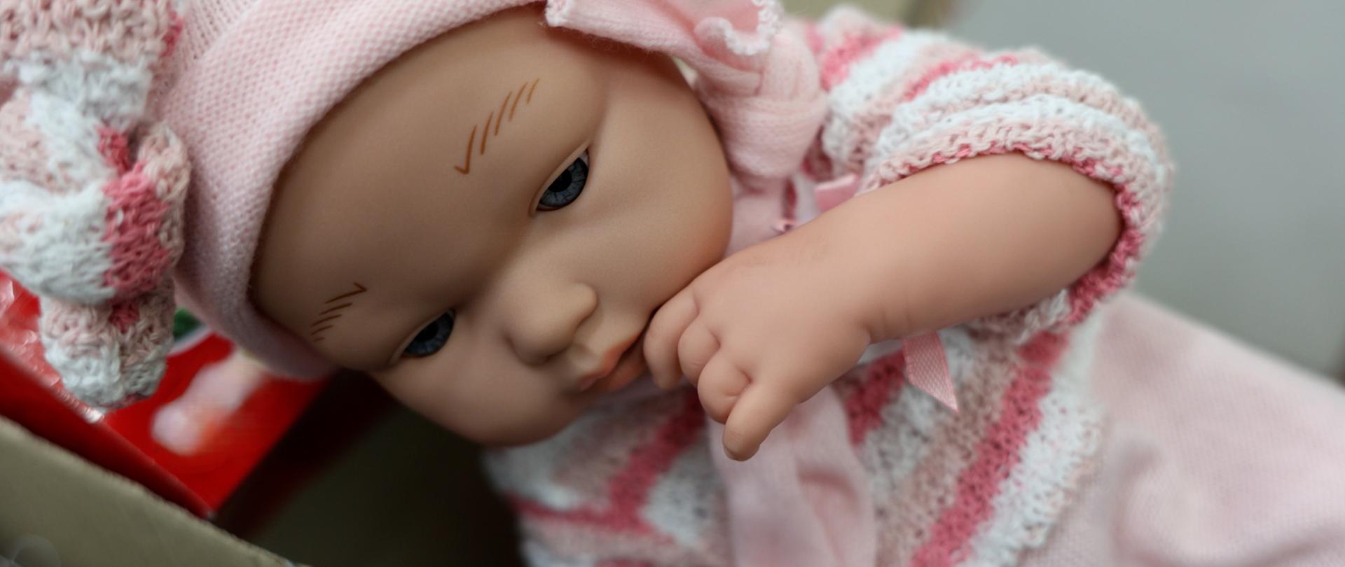 Zbliżenie na lalkę stylizowaną na niemowlę, ubraną w różową czapkę i dzianinowy strój, leżącą w pudełku z ręką przy twarzy.
