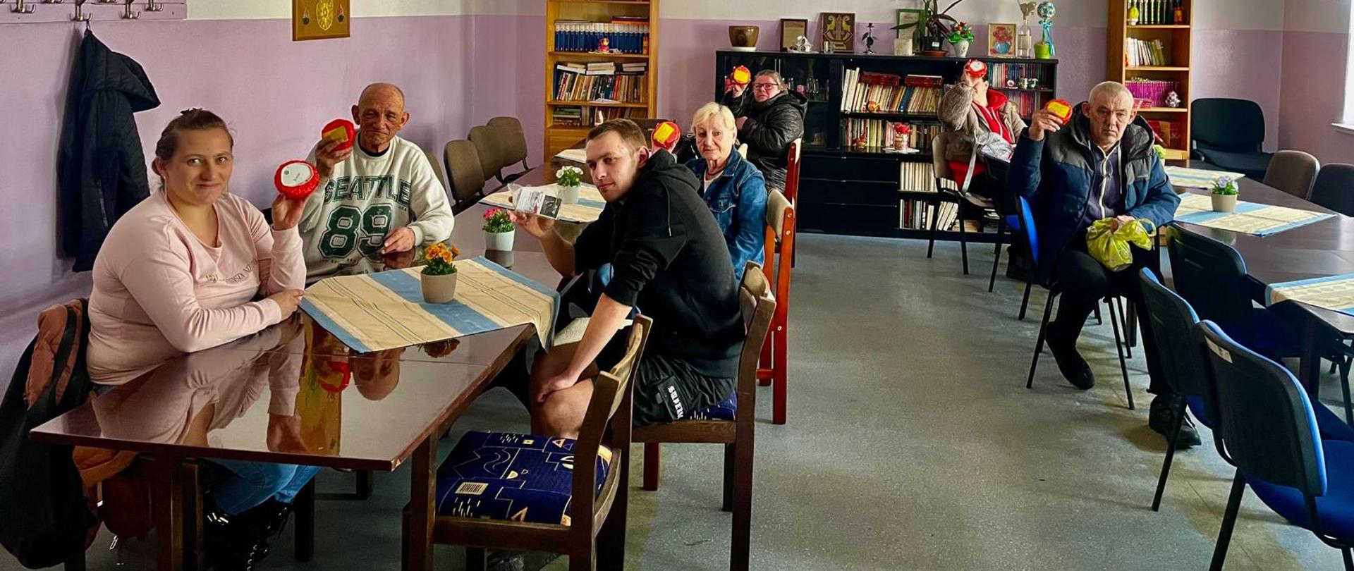 Grupa kilku osób siedzi przy stołach w świetlicy lub sali spotkań. Każda z osób trzyma w ręku czerwony element zestawu czujnika dymu i pokazuje go w stronę aparatu. W tle znajdują się regały z książkami, krzesła oraz dekoracje na ścianach.