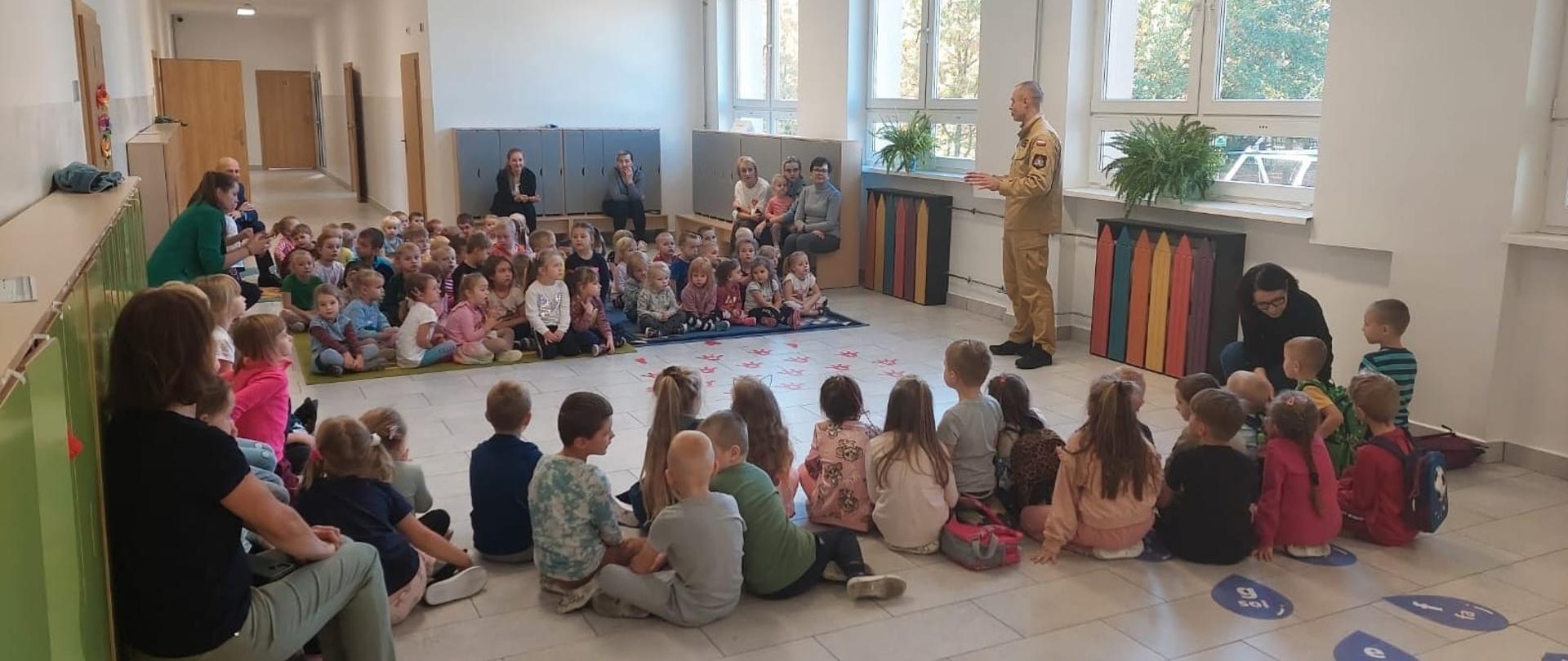 Zdjęcie przedstawia dzieci siedzące na ziemi oraz opiekunów siedzących na ławce, którzy słuchają strażaka.
