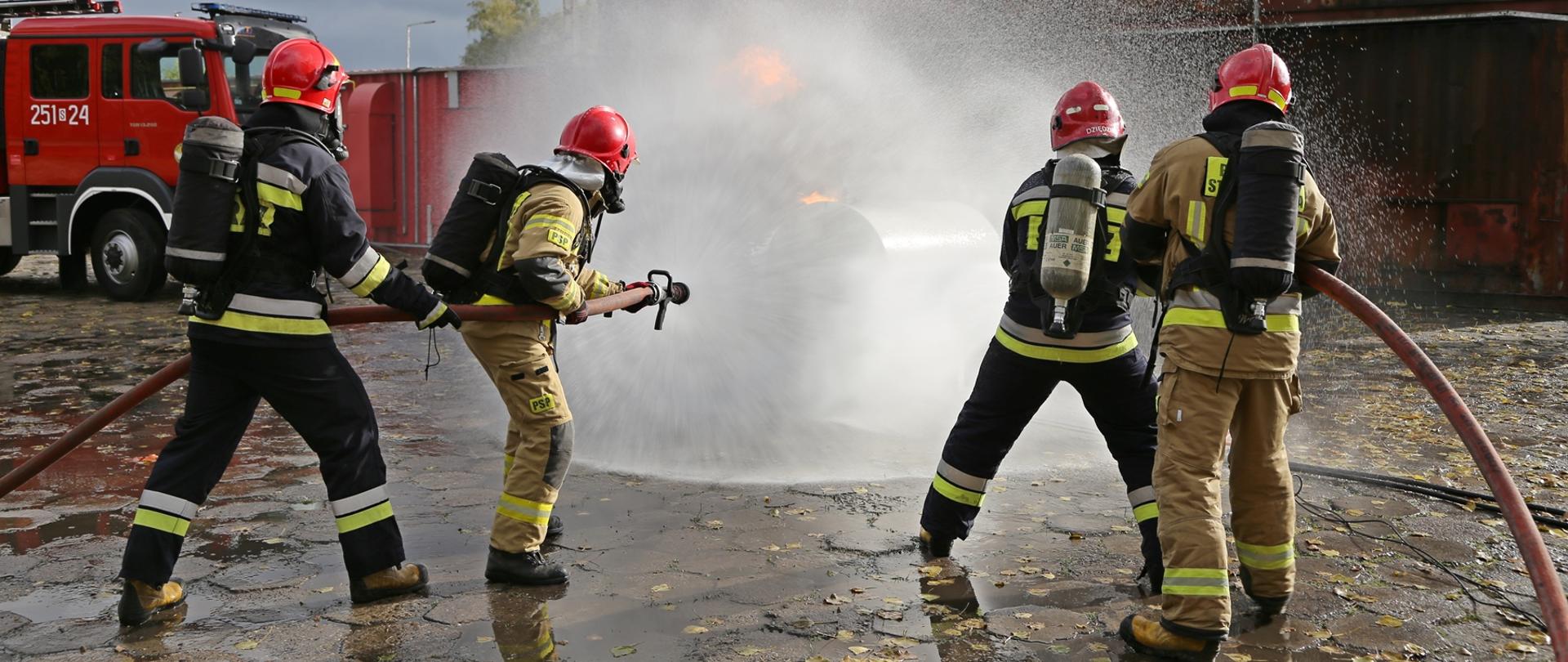 Widok z tyłu. Po lewej stronie dwóch strażaków w umundurowaniu bojowym trzyma wąż gaśniczy, po prawej stronie również dwóch strażaków w umundurowaniu bojowym trzyma wąż gaśniczy. Obie roty kierują strumień wody na źródło ognia. W głębi po lewej widoczny wóz gaśniczy