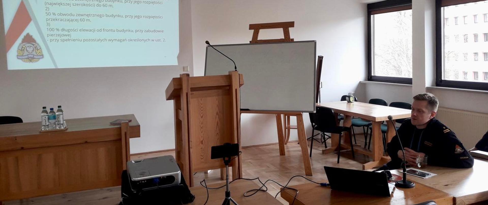 Na zdjęciu widoczny siedzący funkcjonariusz KW PSP Szczecin prowadzący szkolenie na temat przepisów ppoż. W centrum zdjęcia widoczny rzutnik, z którego prezentowana jest prezentacja na ścianie.
W tle okna, mównica, tablica.