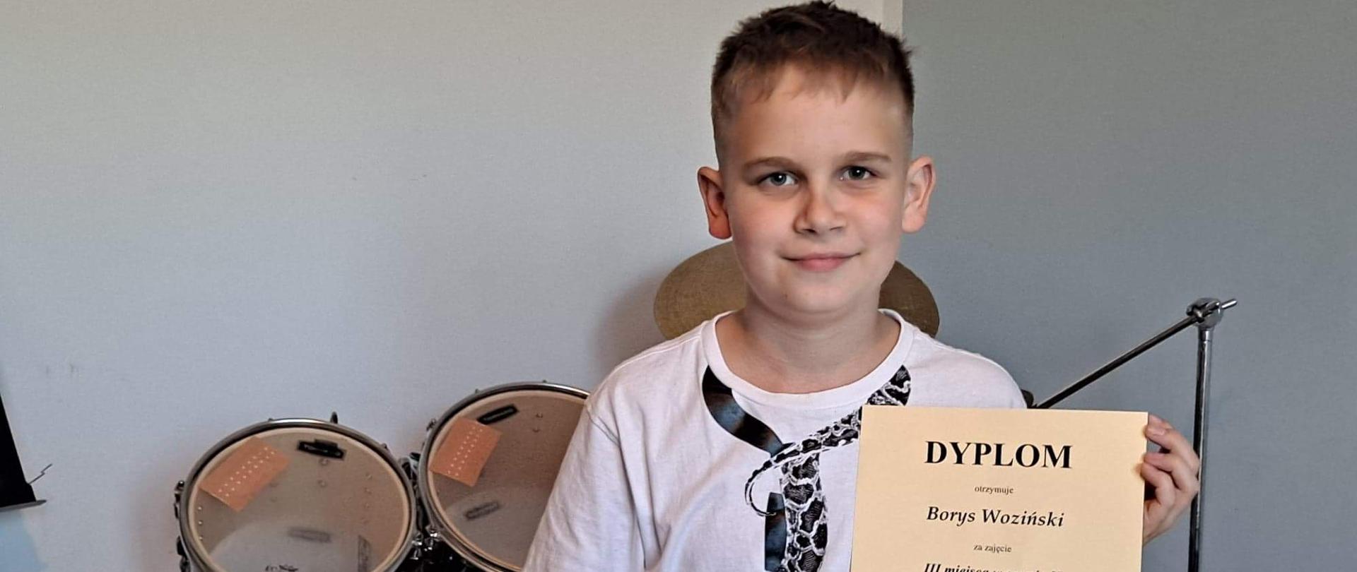 Chłopiec siedzi, w ręku trzyma dyplom, obok zestaw perkusyjny, 