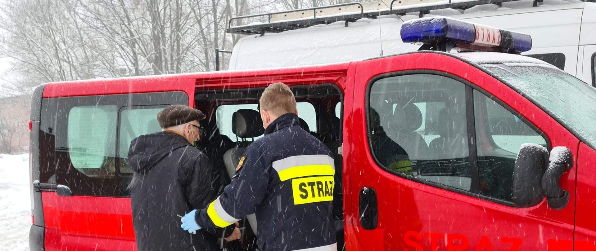 Przed samochodem strażackim typu bus Ochotniczej Straży Pożarnej w Nowem, który ma otwarte drzwi przesuwne, stoi strażak w ubraniu specjalnym i podtrzymuje lewą ręką starszego Pana pod prawe ramię, który przygotowuje się aby wsiąść do środka samochodu.