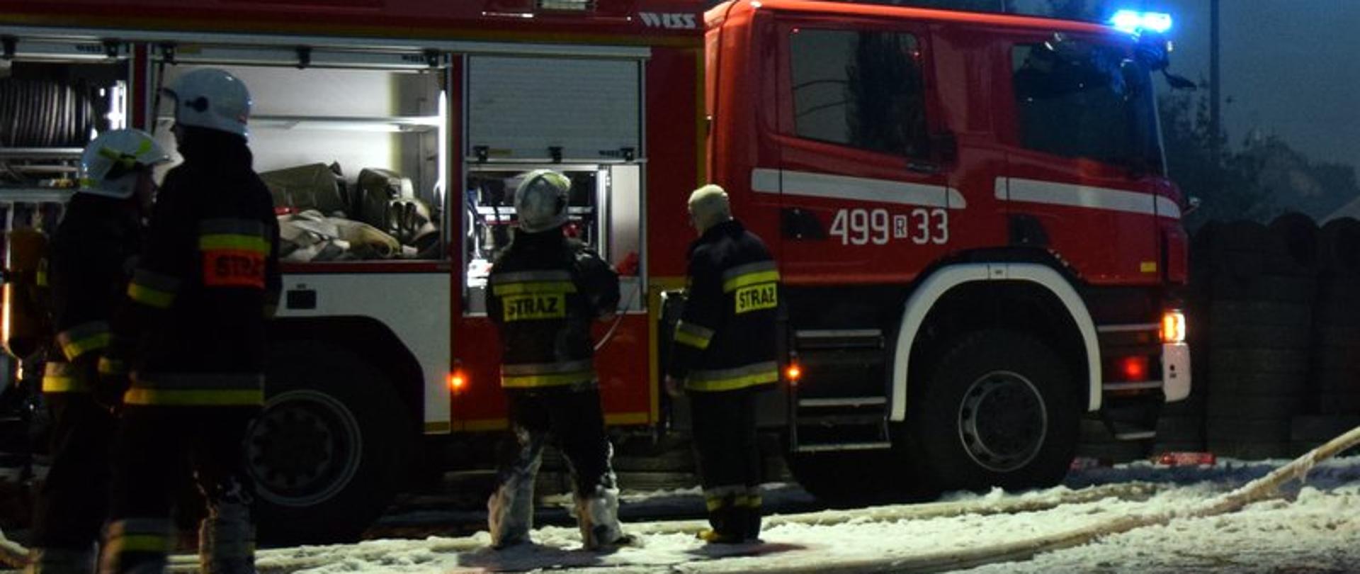 Czterech strażaków stojących na tle samochodu pożarniczego. Zdjęcie zrobione w nocy. Strażacy przygotowują się do gaszenia pożaru zakładając aparaty powietrzne. Zdjęcie ilustracyjne.