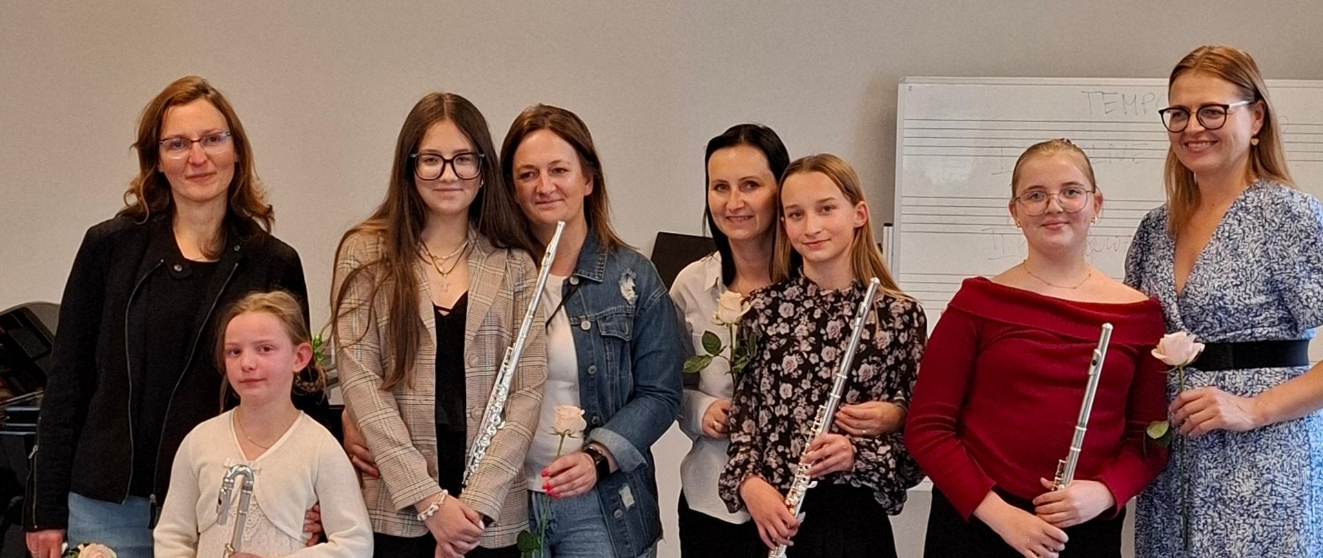 Na zdjęciu widzimy grupę ośmiu osób, w tym dorosłych kobiet i dziewcząt, stojących razem w jasnym pomieszczeniu, przypominającym salę koncertową lub lekcyjną, z instrumentami muzycznymi – fletami. Od lewej strony, stoją dorosła kobieta w ciemnej kurtce i jeansach, trzymająca różę, a obok niej mała dziewczynka w jasnej, koronkowej sukience, trzymająca flet poprzeczny. Następnie, obok, stoi kolejna dorosła kobieta w jasnym swetrze i ciemnych spodniach, a obok niej młodsza kobieta w jeansowej kurtce, również trzymająca różę i flet. Wszystkie patrzą w stronę obiektywu. Pośrodku kadru, w białej bluzce z kwiatowym wzorem, stoi dorosła kobieta, a obok niej dziewczynka w czarnej spódniczce i bluzce w kwiaty, trzymająca flet. Dalej na prawo, stoi dziewczynka w czerwonej bluzce i czarnej spódniczce, trzymająca flet. Na samym końcu, po prawej stronie, w sukience w niebieskie wzory, stoi dorosła kobieta, trzymająca różę.
W tle widać jasne ściany i fragment tablicy nutowej, a także głośnik na ścianie po lewej stronie. Wszystkie osoby uśmiechają się, a atmosfera zdjęcia jest radosna i uroczysta, sugerując zakończenie występu lub lekcji.